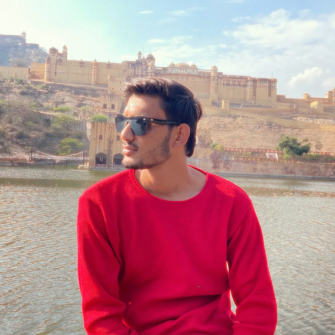 A person in a red sweater and sunglasses beside a lake, with a fort in the background
